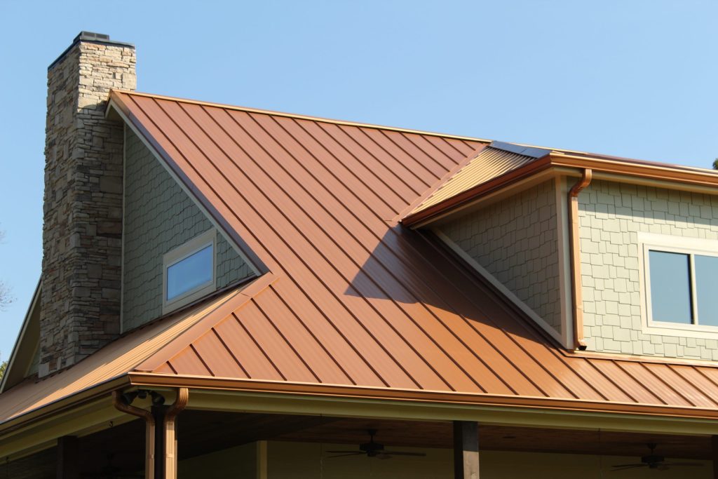 Copper standing seam metal roof panels on a modern home with stone chimney, dormer and matching gutters under clear blue sky.