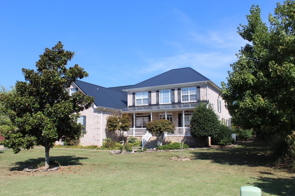 Two-story brick home with exposed fastener metal roof panels, covered front porch, and landscaped yard.