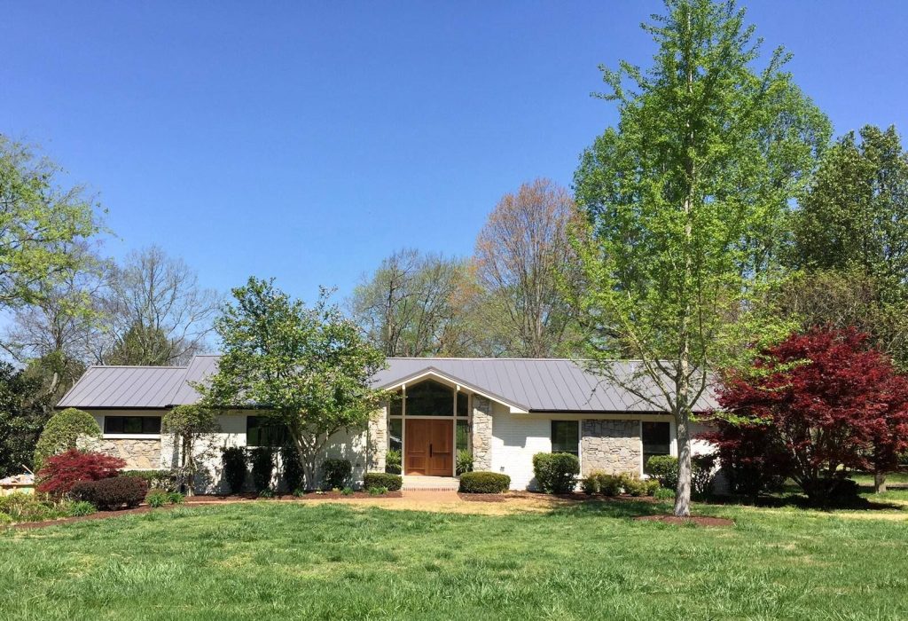 Modern ranch home with gray standing seam metal roof panels, stone and white brick exterior, and landscaped front yard under blue sky.