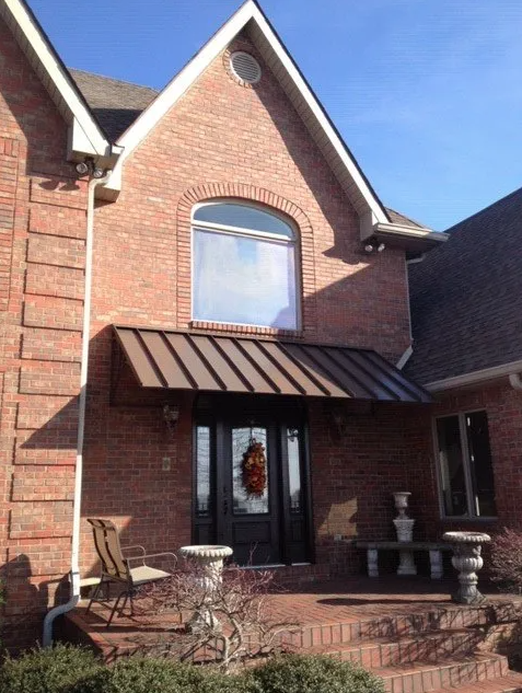 Brick home entry with brown standing seam metal roof awning panel above front door, arched window, and brick steps leading to porch.