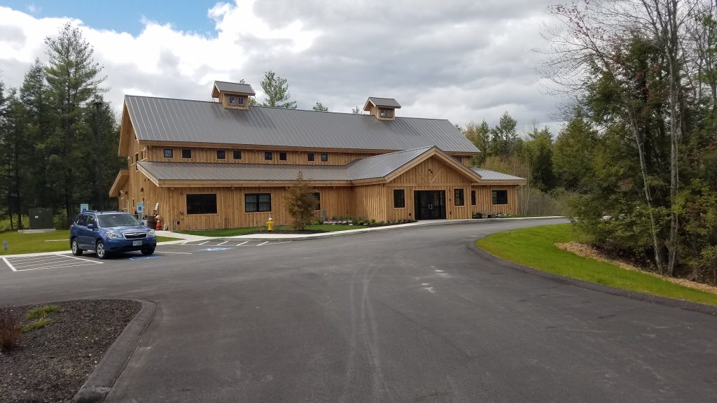 Wood-framed commercial building with gray standing seam metal roof panels, cupolas, and covered entry, surrounded by trees and parking lot.